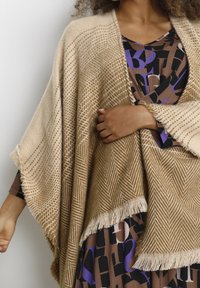 Beige sjal med hundtands- och fiskbensmönster, med fransade kanter. Bärs över en mönstrad brun klänning med svarta och lila accenter.