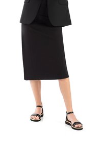 Schwarzer Bleistiftrock kombiniert mit schwarz-weißen Sandalen mit zwei Riemen, die über eine dicke Sohle und einen Knöchelriemen verfügen, vor einem weißen Hintergrund präsentiert.