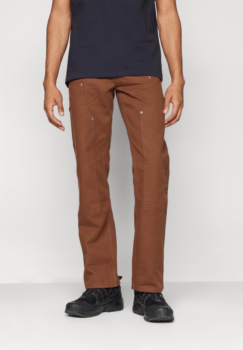 Brown pants with a straight-leg fit, made of a durable fabric. Features visible stitching and metal rivets. Paired with black shoes.