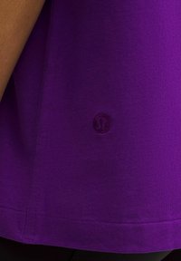 Violeta sporta krekls, kas izgatavots no gludas, elastīgas auduma ar apaļu kakla izgriezumu un mazu izšuvumu logo apakšējā labajā stūrī.