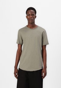 Olivgrünes T-Shirt mit kurzen Ärmeln, rundem Ausschnitt, leicht abgerundetem Saum, rohen Kanten und einem kleinen Logo an der Seite.