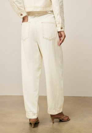 Naine, kes kannab kreemjaid kõrge vöökohaga laiade pükse tagataskutega ja sobivaid jopesid, seisab ühe käega taskus ja tal on jalas beežid kontsaga sandaalid.