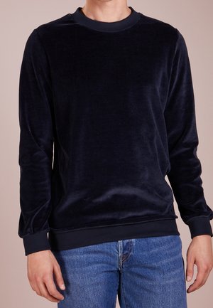 Mand iført en sort fløjlsblød crewneck sweatshirt med lange ærmer og blå denimjeans, stående mod en ensfarvet baggrund.