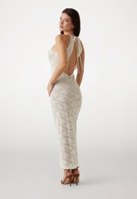 Beiges Strickkleid mit offenem Rückendesign, langer Silhouette, strukturiertem Muster und verstellbaren Bindebändern. Kombiniert mit braunen High-Heels.