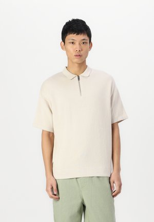 Polo shirt beige clair à manches courtes avec une fermeture éclair sur le devant et un col structuré, associé à un pantalon vert clair. Texture lisse.