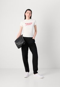 Valkoinen lyhythihainen t-paita, jossa on punainen "tommy jeans" -teksti, yhdistettynä mustiin korkeavyötäröisiin housuihin ja valkoisiin tennareihin, pitäen käsissä mustaa clutch-pussia.