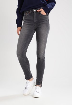 Vaqueros de mezclilla gris de talle alto con corte ajustado, detalles desgastados y dobladillos deshilachados en los tobillos, combinados con zapatillas blancas.