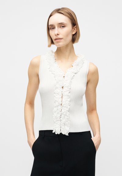 Γυναίκα με λευκή αμάνικη μπλούζα με βολάν μπροστά και μαύρο παντελόνι, που στέκεται μπροστά σε απλό ανοιχτό φόντο.