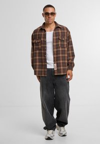 Brown plaid flannel shirt with two chest pockets, buttoned front, paired with loose-fitting dark gray pants and textured sneakers.