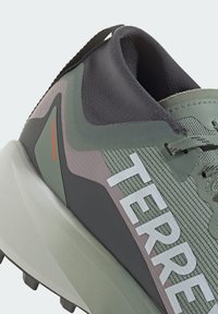 Baskets de sport vertes et grises avec tige en mesh, semelle épaisse, talon prononcé et marque blanche. Accents rouges sur le côté. Surface texturée.