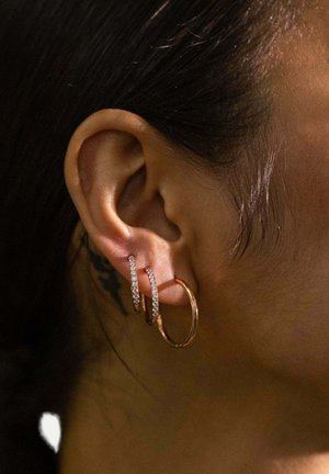 Ohr mit drei Creolen: zwei kleine silberne Creolen mit Diamanten und eine größere schlichte goldene Creole, vor dunklem Haar und Haut.