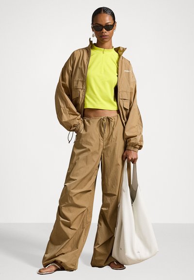 Brūna viegla jaka, neona zaļa crop top un atbilstošas plašas bikses. Balta hobo soma un tumšas saulesbrilles noslēdz ansambli.