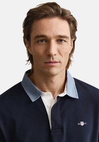 Donkerblauw poloshirt met een lichtblauwe kraag, witte knoopsluiting en GANT-logo op de borst. Gladde textuur, klassieke pasvorm.