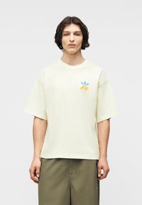 Νεαρός με μακριά μέχρι τους ώμους σκούρα μαλλιά, φορώντας μια άνετη λευκή μπλούζα με μικρό μπλε και κίτρινο λογότυπο, και χακί παντελόνι.