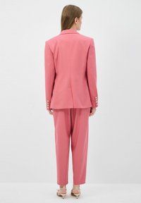 Vaaleanpunainen puku, jossa on räätälöity bleiseri, jossa on olkatyynyt ja kultanapit, yhdistettynä vastaaviin kapeneviin housuihin. Silkkinen kangas ja jämäkkä muotoilu.