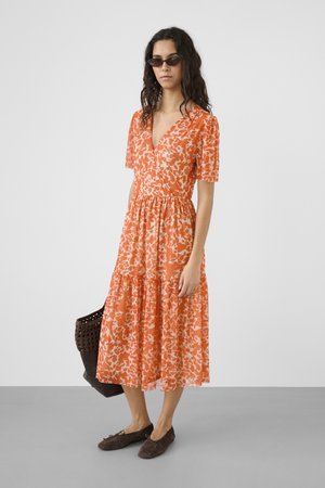 Frau mit langen lockigen Haaren, die ein orange-weißes Midikleid mit Blumenmuster trägt, braune Ballerinas, Sonnenbrille und eine dunkle gewebte Handtasche in der Hand hält.