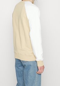 Suéter de algodón con cuerpo beige y mangas blancas, puños y dobladillo acanalados. Textura suave con detalles de costura visibles.