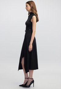 Schwarzes ärmelloses Kleid mit hohem Kragen, tailliertem Oberteil und fließendem Rock. Es hat einen seitlichen Schlitz und einen schwarzen Gürtel in der Taille.