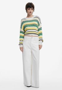 Großer Strickpullover mit gelben, grünen und schwarzen Streifen, kombiniert mit weißen weiten Hosen. Der Pullover hat lange Ärmel und ein kurzes Design.