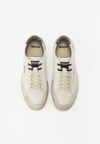 Hvite sneakers med perforert tå, beige teksturerte detaljer, sorte snører, rutet krage og gummigummisåle. Merkevare på siden og tungen.