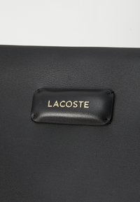Crna kožna torba s teksturiranom površinom i zlatno otisnutim Lacoste logotipom na pravokutnom kožnom zakrpu.