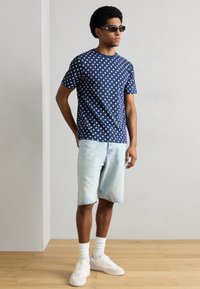 Μπλε ναυτικό t-shirt με λευκές πουά βούλες, συνδυασμένο με ανοιχτόχρωμο τζιν σορτς. Το μοντέλο φορά γυαλιά ηλίου και λευκά αθλητικά παπούτσια με υφή στη σόλα.