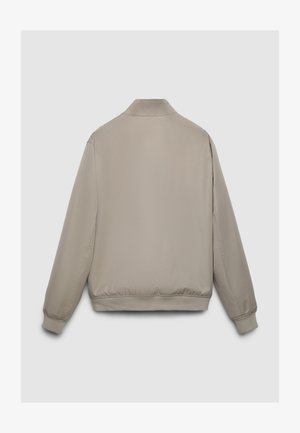 Beige Bomberjacke mit glatter Textur, ausgestattet mit einem Stehkragen, elastischen Bündchen und Saum, die ein minimalistisches Design zeigen.