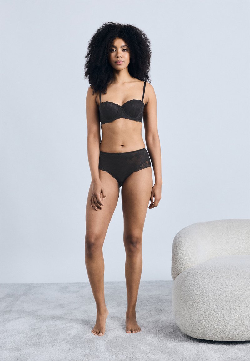 Μαύρο δαντελένιο σουτιέν και ταιριαστόι ψηλόμεσοι σλιπ, με διάφανα στοιχεία. Η μοντέλο στέκεται σε φωτεινή γκρι υφή επιφάνειας, δίπλα σε λευκό καναπέ.