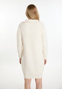 Gebreide crèmekleurige trui-jurk, halflang met een ribbelstructuur, brede kraag en lange mouwen. Eenvoudig ontwerp zonder patronen of accenten.