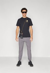 Μαύρο μπλουζάκι με χρυσό γραφικό, γκρι παντελόνι και μαύρα αθλητικά παπούτσια. Μια μικρή θήκη για το τηλέφωνο, προσαρτημένη με αλυσίδα, κρέμεται διαγώνια στο σώμα.