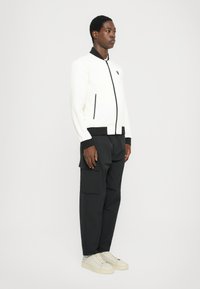 Jachetă bomber albă cu buzunare cu fermoar și accente negre, asociată cu pantaloni cargo negri și teniși crem. Textură netedă, design modern.