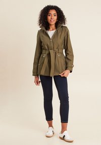 Olijfgroene, gebelt jasje met ritssluiting en zijzakken, gecombineerd met marineblauwe slim fit broek en witte sneakers. Gestructureerde stof en verstelbare kraag.