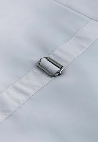 Metallspänne med dragkedja fäster en ljusgrå justerbar tygrem på en slät grå textilyta.