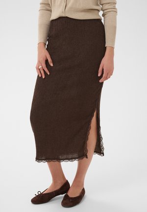 Femme portant une jupe midi marron texturée avec bordure en dentelle sur le côté et une fente, associée à des ballerines marron foncé et un haut beige à manches longues.
