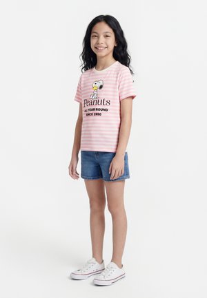 Smilende pige med langt sort hår iført en lyserød stribet Peanuts T-shirt, denimshorts og hvide sneakers, stående mod en hvid baggrund.