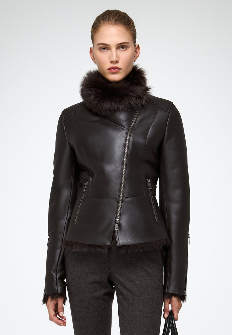 Donna che indossa una giacca di pelle nera con collo e polsini di pelliccia, abbinata a pantaloni grigio scuro, mentre tiene una borsetta nera.