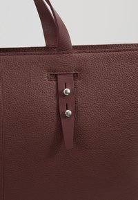 Détail d'un sac cabas en cuir texturé marron foncé montrant la sangle avec deux clous argentés et des bords cousus.