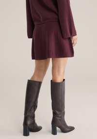 Knielange braune Lederstiefel mit Blockabsatz, kombiniert mit einem kastanienbraunen gestrickten Rock und Pullover, die einen lässigen Lagenlook präsentieren.