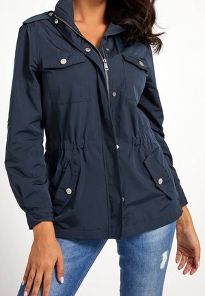 Kvinde iført en marineblå lynlåstrøje med fire lomme med knapper og en hætte, kombineret med slidte blå jeans.