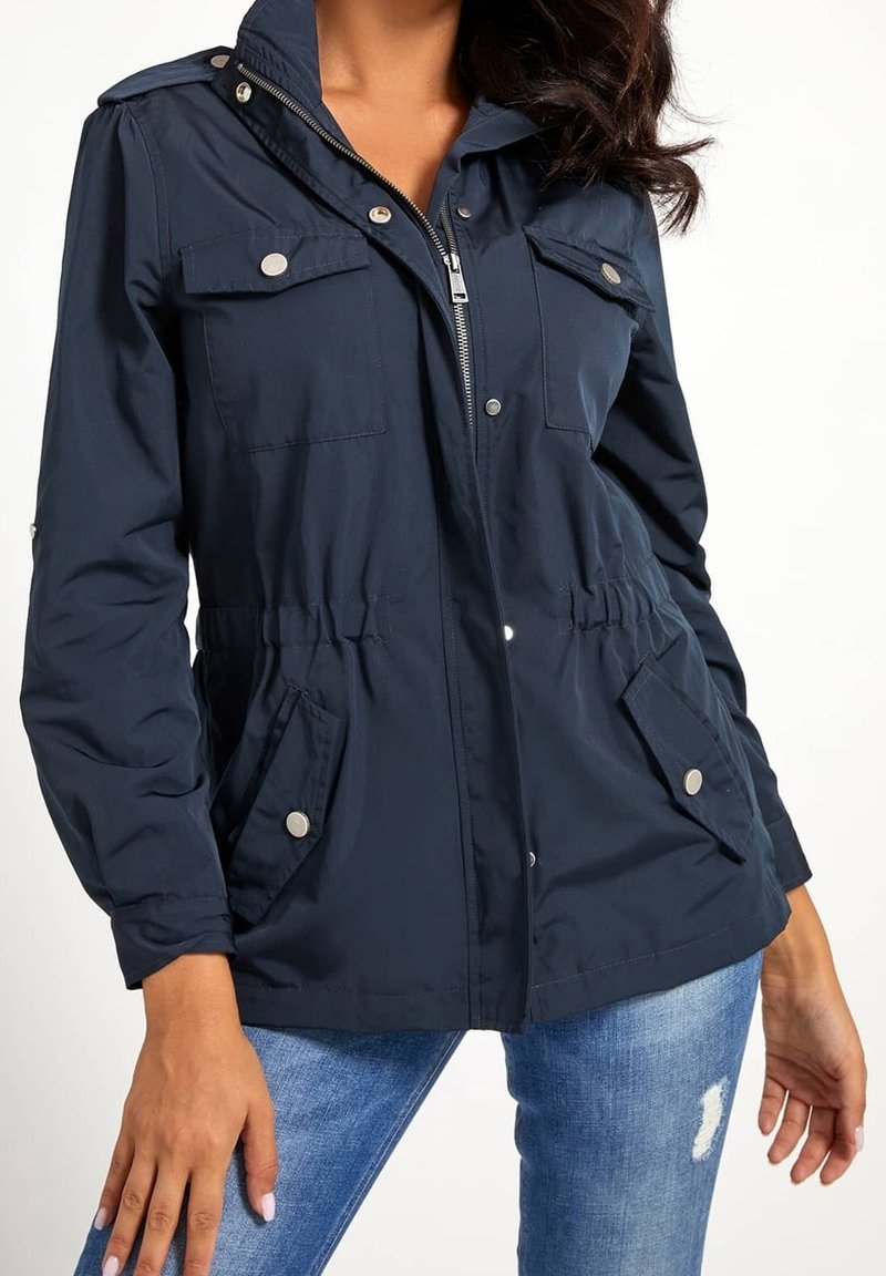 Femme portant une veste zippée bleu marine avec quatre poches boutonnées et une capuche, associée à un jean bleu déchiré.
