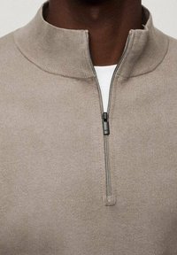 Gros plan d'une personne portant un pull beige à demi-fermeture éclair sur une chemise blanche, montrant la fermeture éclair partiellement ouverte au niveau du col.