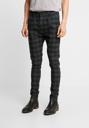 Mann trägt schlanke dunkelgraue karierte Hose und schwarze Leder-Stiefeletten, steht auf hellem einfarbigem Hintergrund.