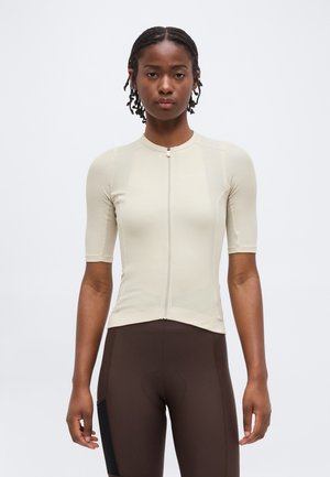 Femeie de culoare cu părul împletit, purtând un tricou de ciclism bej cu mâneci scurte și fermoar și pantaloni scurți căptușiți de culoare maro închis pe fundal simplu.