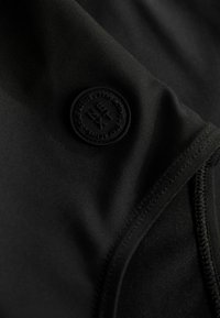 Černá sportovní tkanina s hladkým povrchem a kulatou vyvýšenou černou gumovou nášivkou s textem "NEXT." Viditelné prošívání podél okrajů.