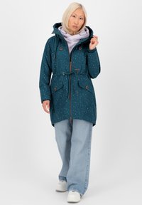 Giacca verde acqua con cappuccio, motivo a pois multicolore, vita con cintura e tasche frontali. Abbinata a jeans in denim chiaro larghi e sneakers bianche.