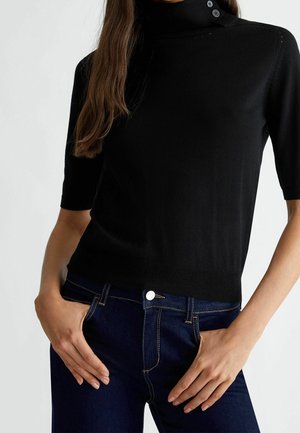 Femme portant un pull à col roulé noir à manches courtes avec des détails de boutons et un jean en denim bleu foncé, les mains reposant près des poches.