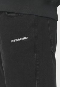 Musta farkut slim fit -mallilla, joissa on vasemmassa reidessä valkoiseksi brodeerattu "PEGADOR" -logo. Silkkinen rakenne, minimaalinen ompelutieto.