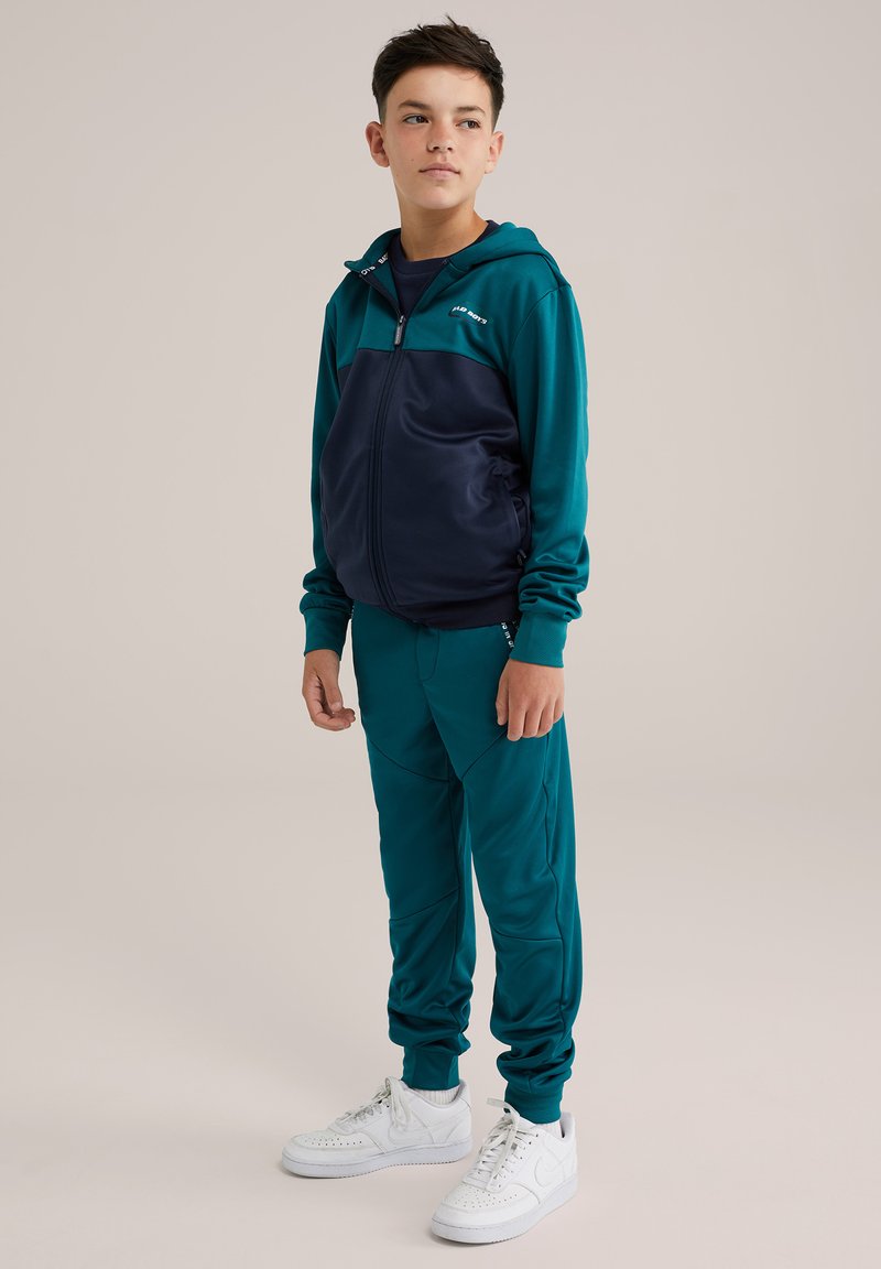 Teal och marinblå träningsdräkt med zip-up huvtröja, som har en tvåfärgad design och slät textur. Matchas med vita sneakers.