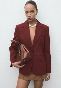 Vrouw in een dieprode blazer en een bruine minirok, met een bruin leren handtasje,accessoires van een parelketting en gouden ooghangers.