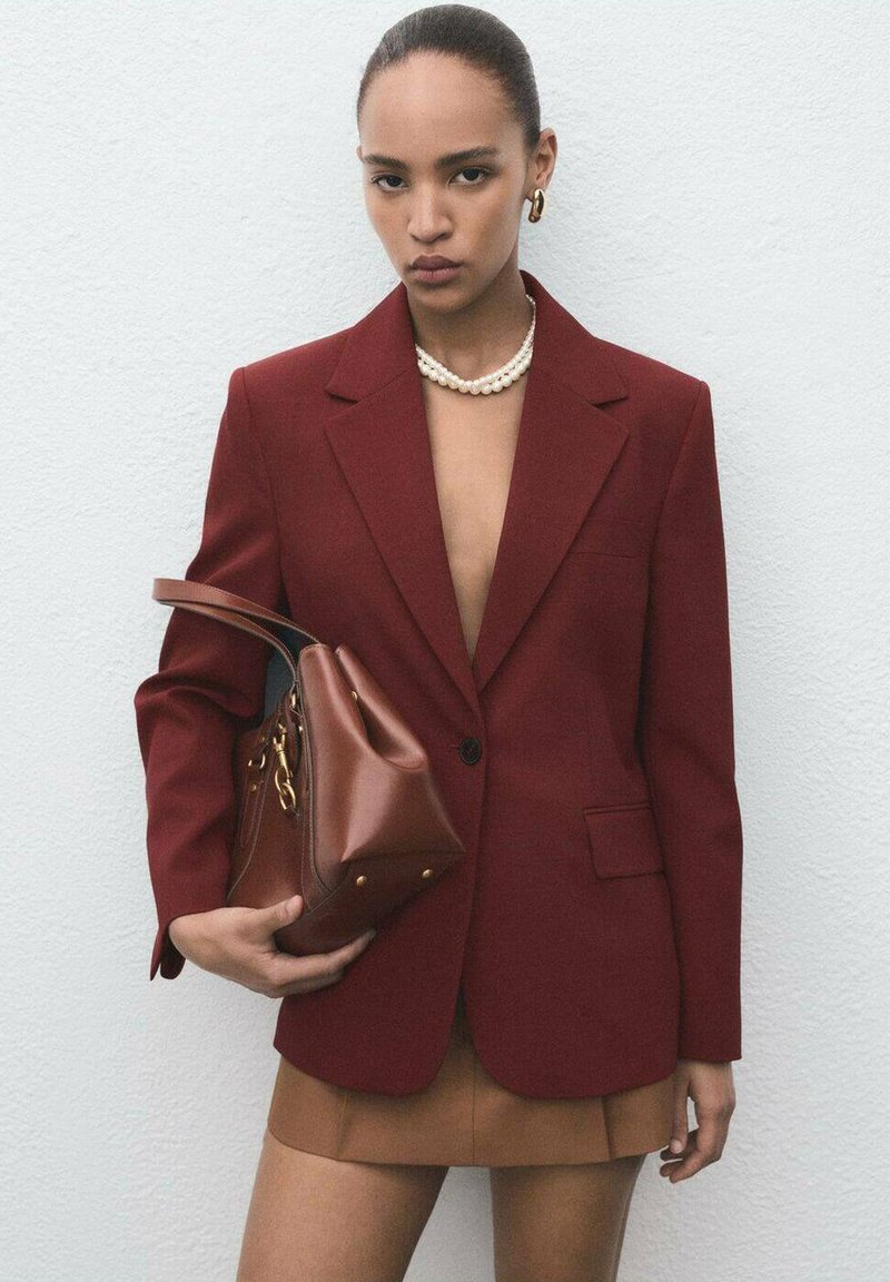 Vrouw in een dieprode blazer en een bruine minirok, met een bruin leren handtasje,accessoires van een parelketting en gouden ooghangers.